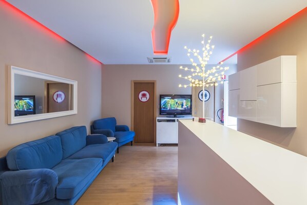 A modern hotel lobby with a blue sofa, a reception desk, and two televisions. - Residencial Vila Nova (Lisbon)