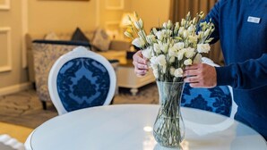 Eine Person hält eine Vase mit weißen Blumen auf einem runden Tisch in einem Raum. Im Hintergrund sind ein stuhl mit blau-weißer Musterung und eine Couch zu sehen.