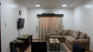 A living room with a beige sofa, a television mounted on the wall, a small table with a tablecloth, and a window with curtains. - Gulf Park Hotel Apartment (Dammam)