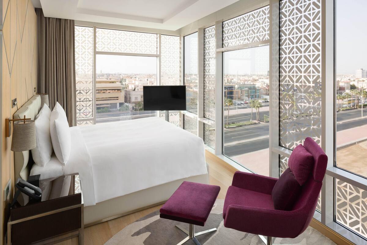 Ein Hotelzimmer mit einem Bett, einem Fernseher, einem violetten Sessel und großen Fenstern mit Blick auf die Stadt.