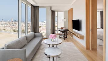 Una habitación de hotel moderna con un ventanal grande, un sofá, una mesa redonda con un florero, un televisor montado en la pared y un armario de madera.
