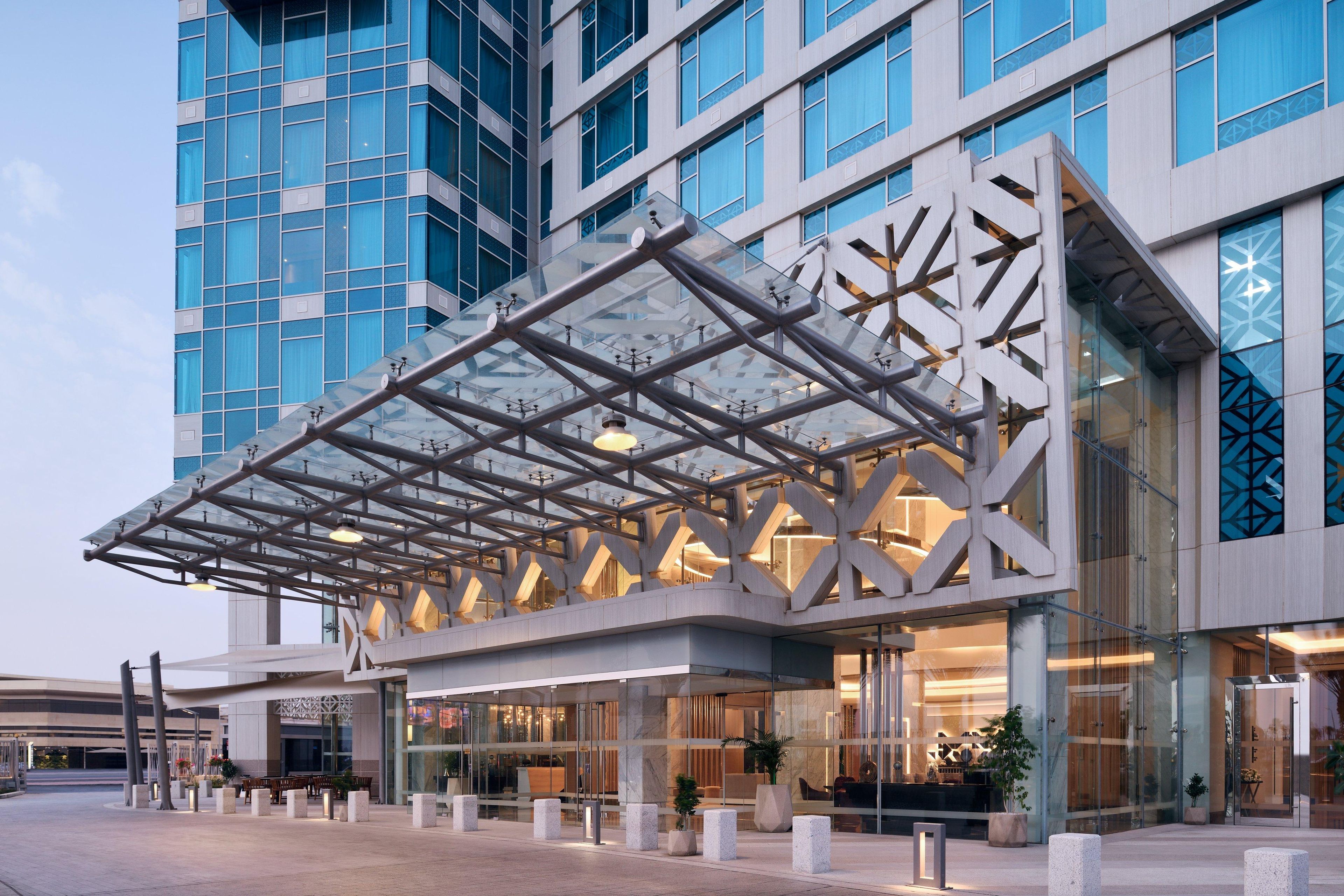 A modern hotel with a glass facade, a distinctive geometric entrance canopy, and a spacious outdoor area with seating.