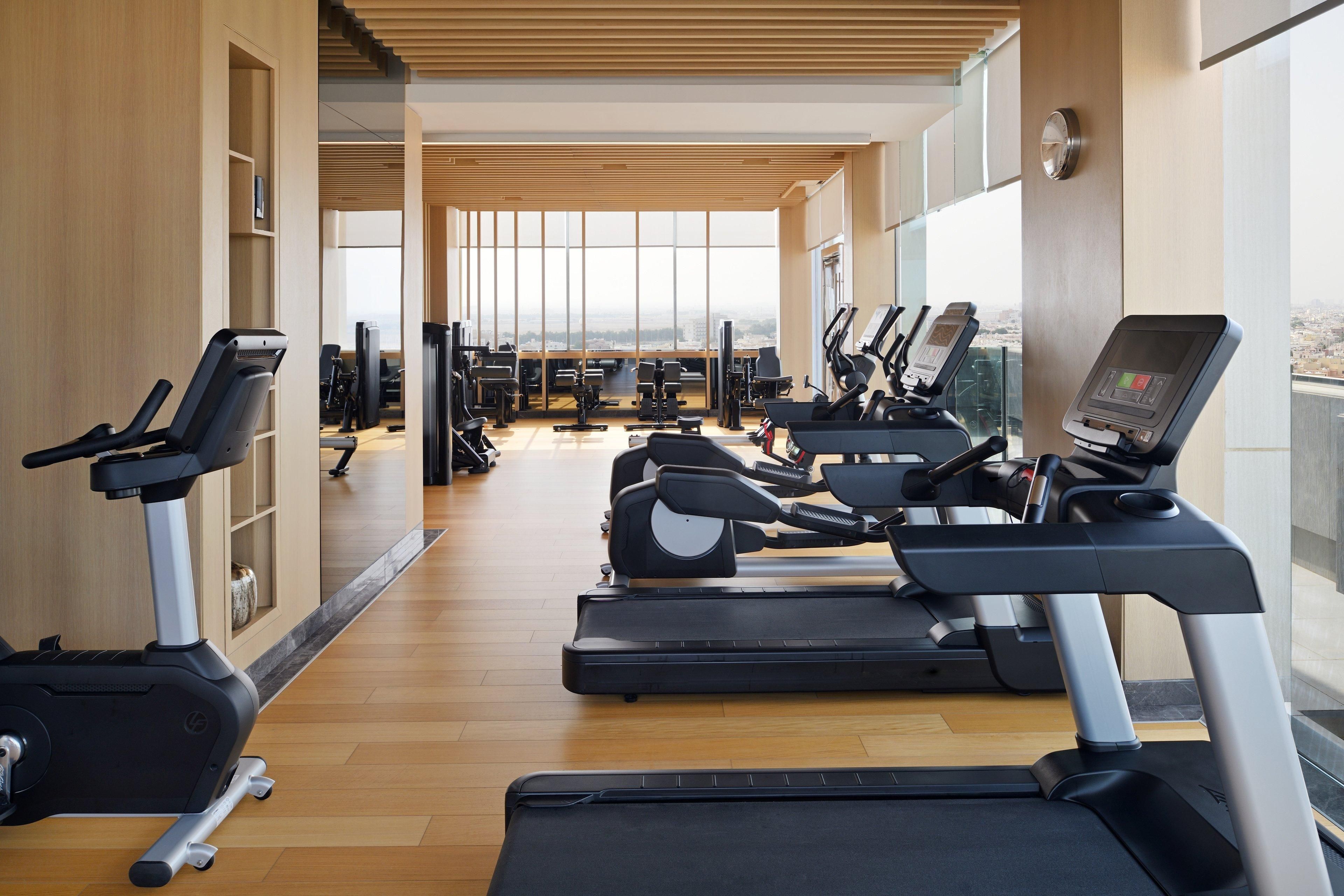 A gym with treadmills, exercise bikes, and weight machines, large windows, and a wooden floor.