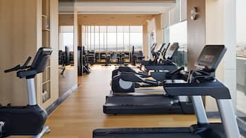 A gym with treadmills, exercise bikes, and weight machines, large windows, and a wooden floor.