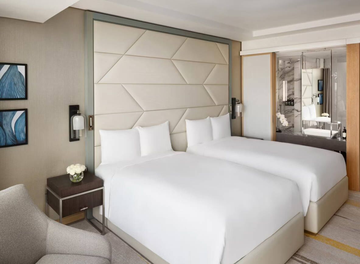 Une chambre d’hôtel avec deux lits, une table de chevet ornée d’un vase de fleurs, une chaise et un grand miroir.