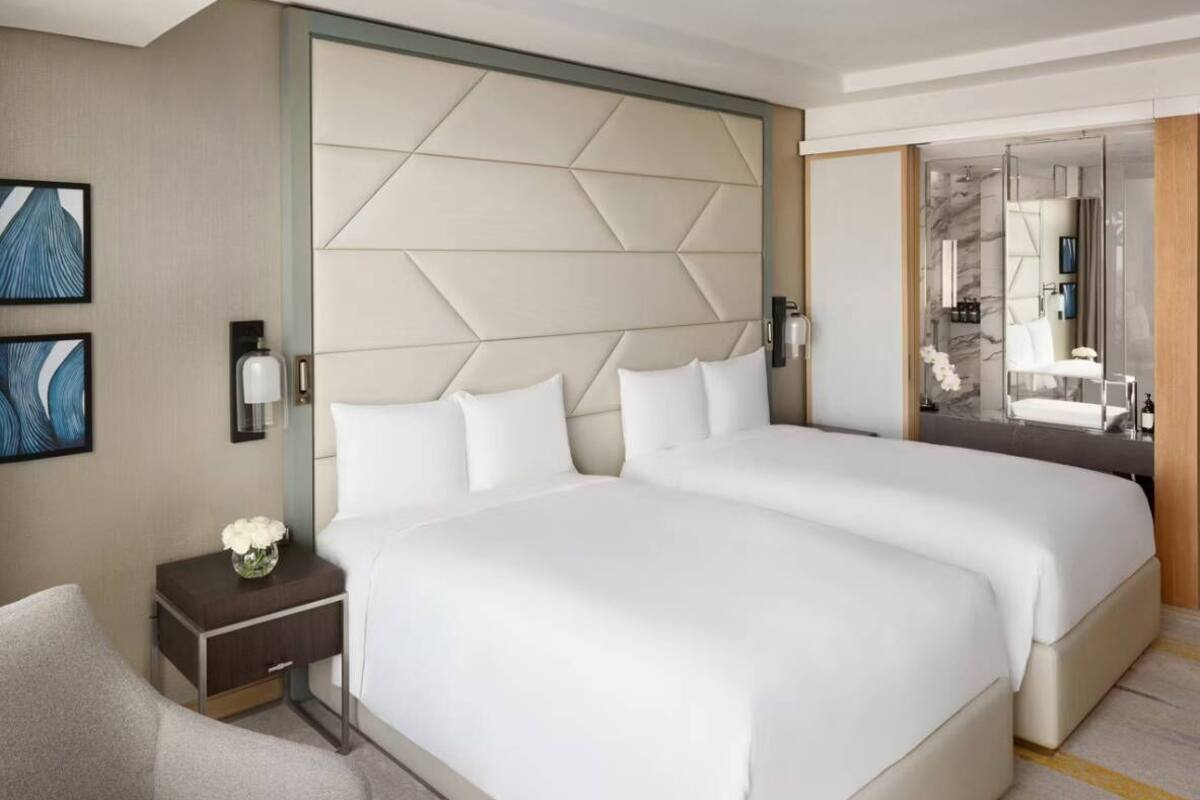 Ein Hotelzimmer mit zwei Betten, einem Nachttisch mit Blumenvase, einem Sessel und einem großen Spiegel.