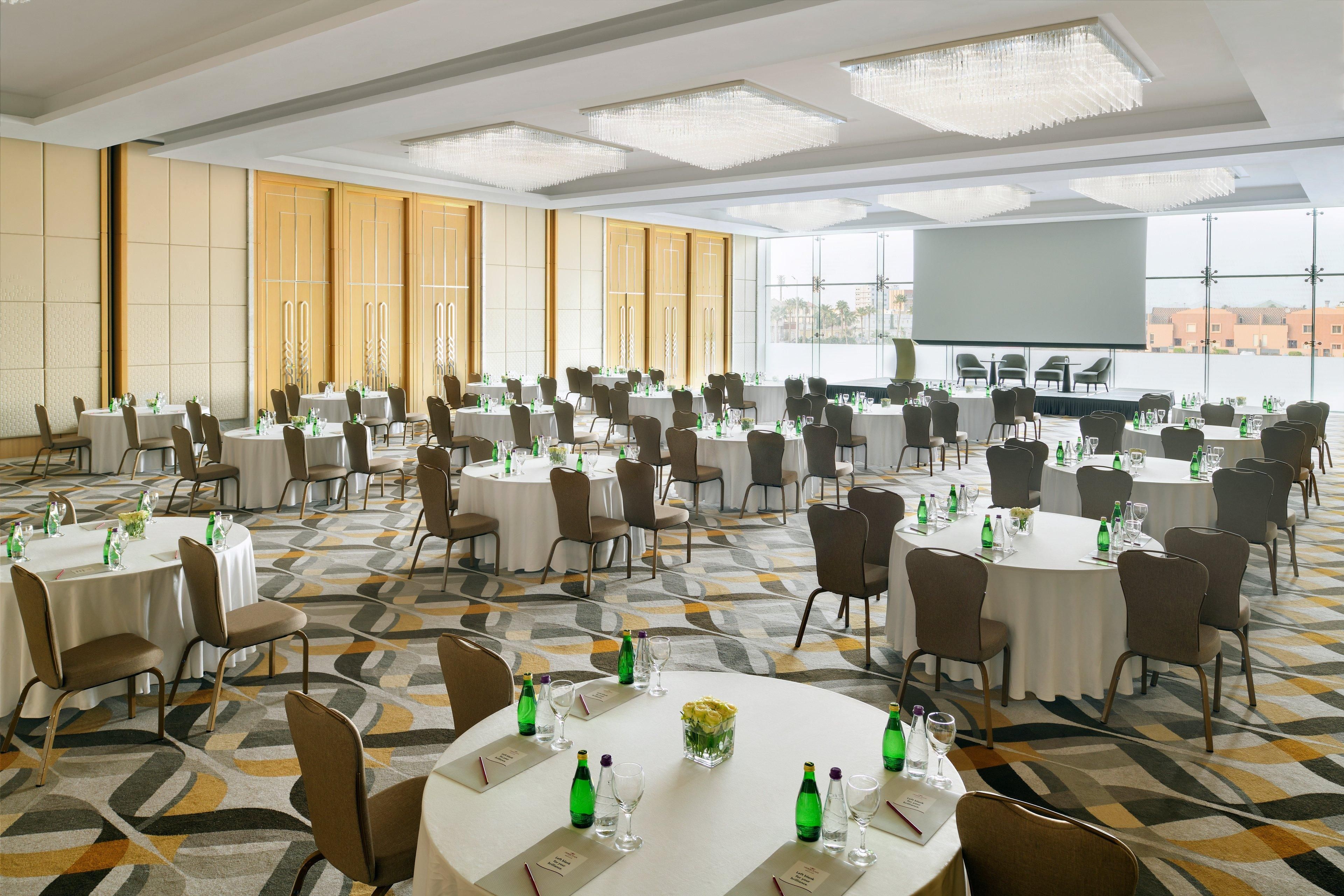 A large conference room with multiple round tables, chairs, and a large screen.
