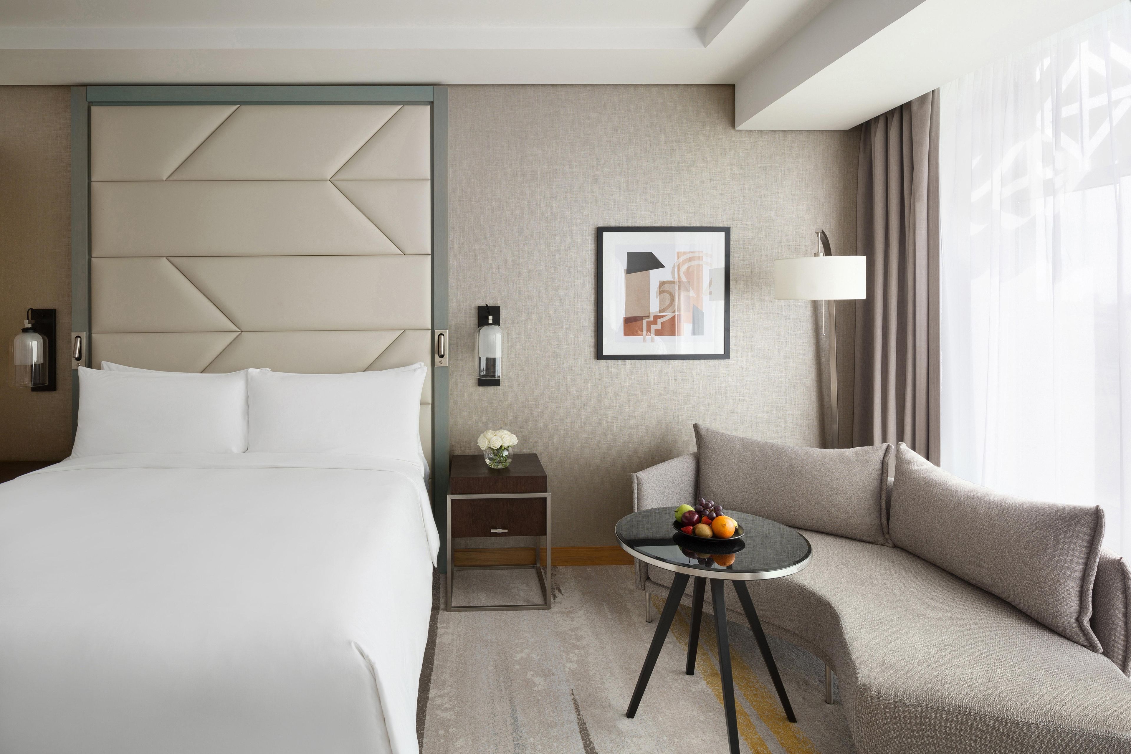 A hotel room with a bed, a sofa, a small table with fruit, a nightstand, a lamp, and a framed picture on the wall.
