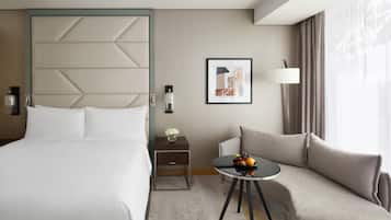 A hotel room with a bed, a sofa, a small table with fruit, a nightstand, a lamp, and a framed picture on the wall.