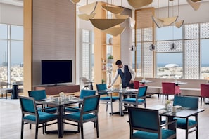 A modern dining area with multiple tables and chairs, a large flat-screen TV, and a view of the cityscape through large windows.