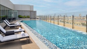 Ein Infinity-Pool auf dem Dach mit Liegestühlen, einer Glasbrüstung und Blick auf die Stadt.