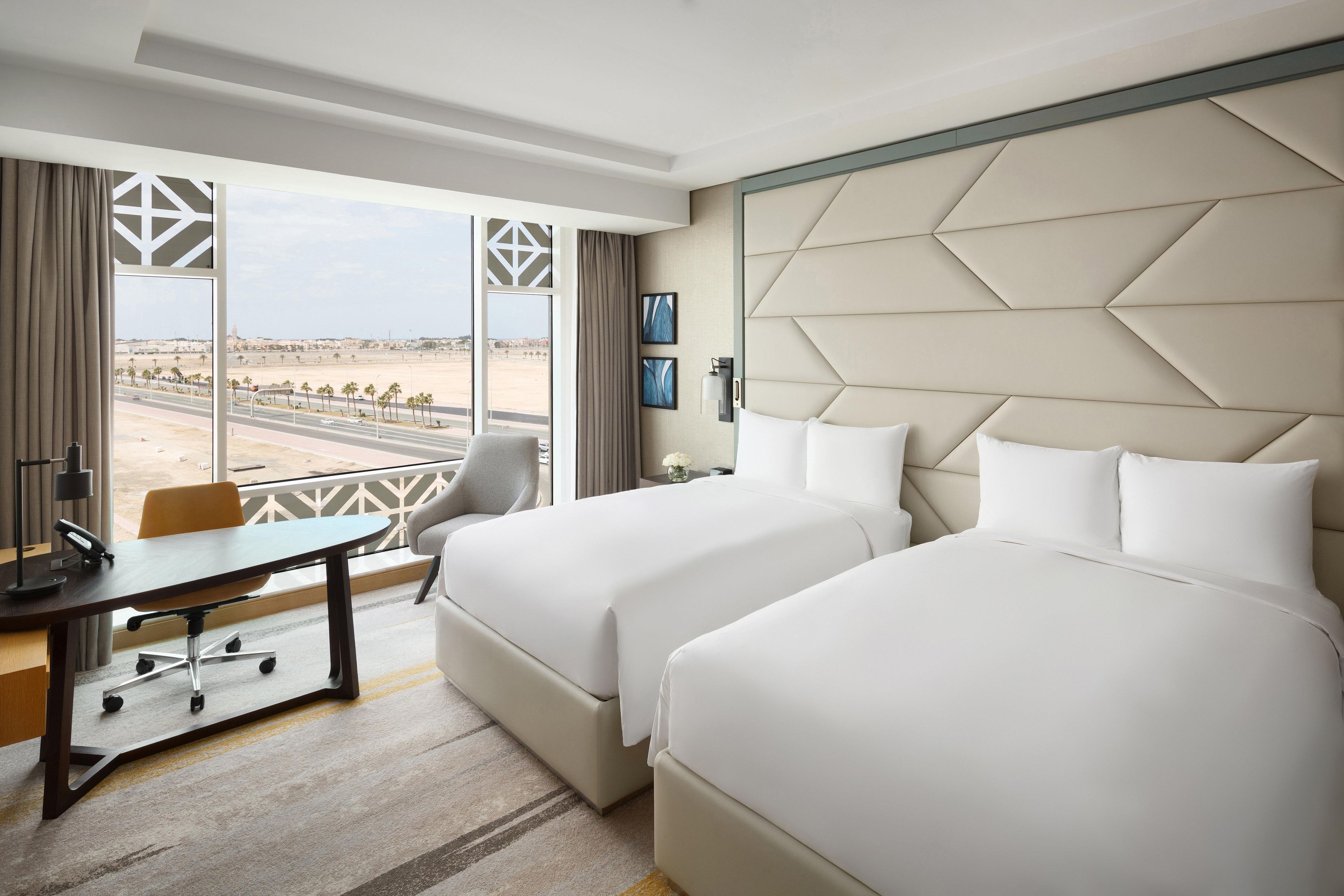 A hotel room with two beds, a desk, and a view of a desert landscape.