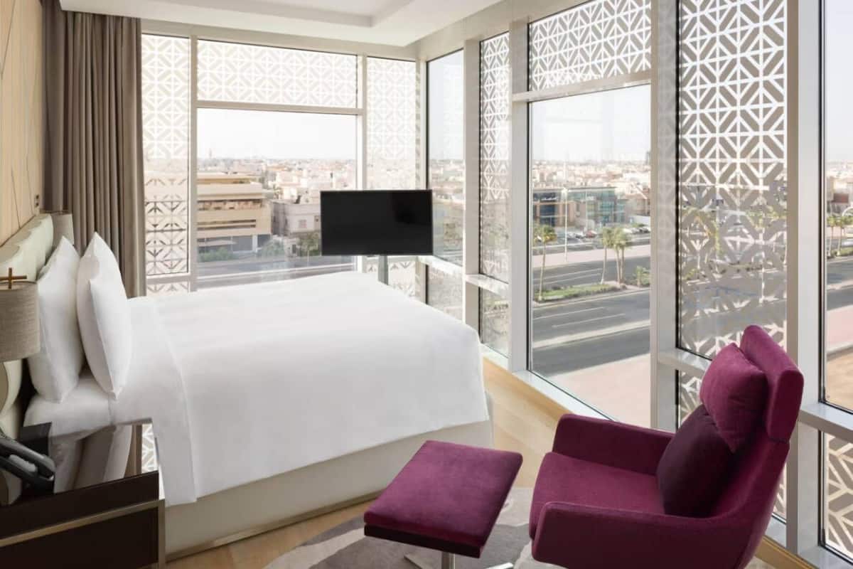 Ein modernes Hotelzimmer mit einem großen Bett, einem Fernseher, einem violetten Sessel und großen Fenstern mit Blick auf die Stadt.