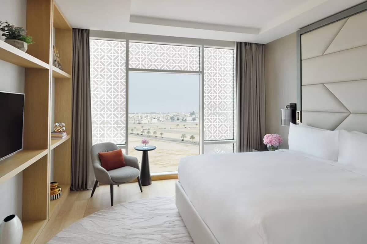 Ein Hotelzimmer mit einem großen Bett, einem Fernseher, einem Sessel, einem kleinen Tisch und Blick auf die Stadt durch bunte Vorhänge.