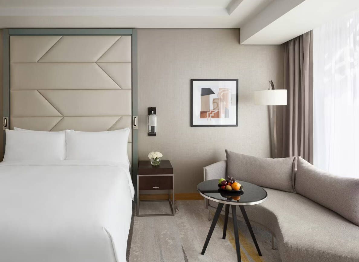 Une chambre d’hôtel avec un lit, une table de chevet, une table avec des fruits, un canapé, une lampe et un tableau encadré au mur.