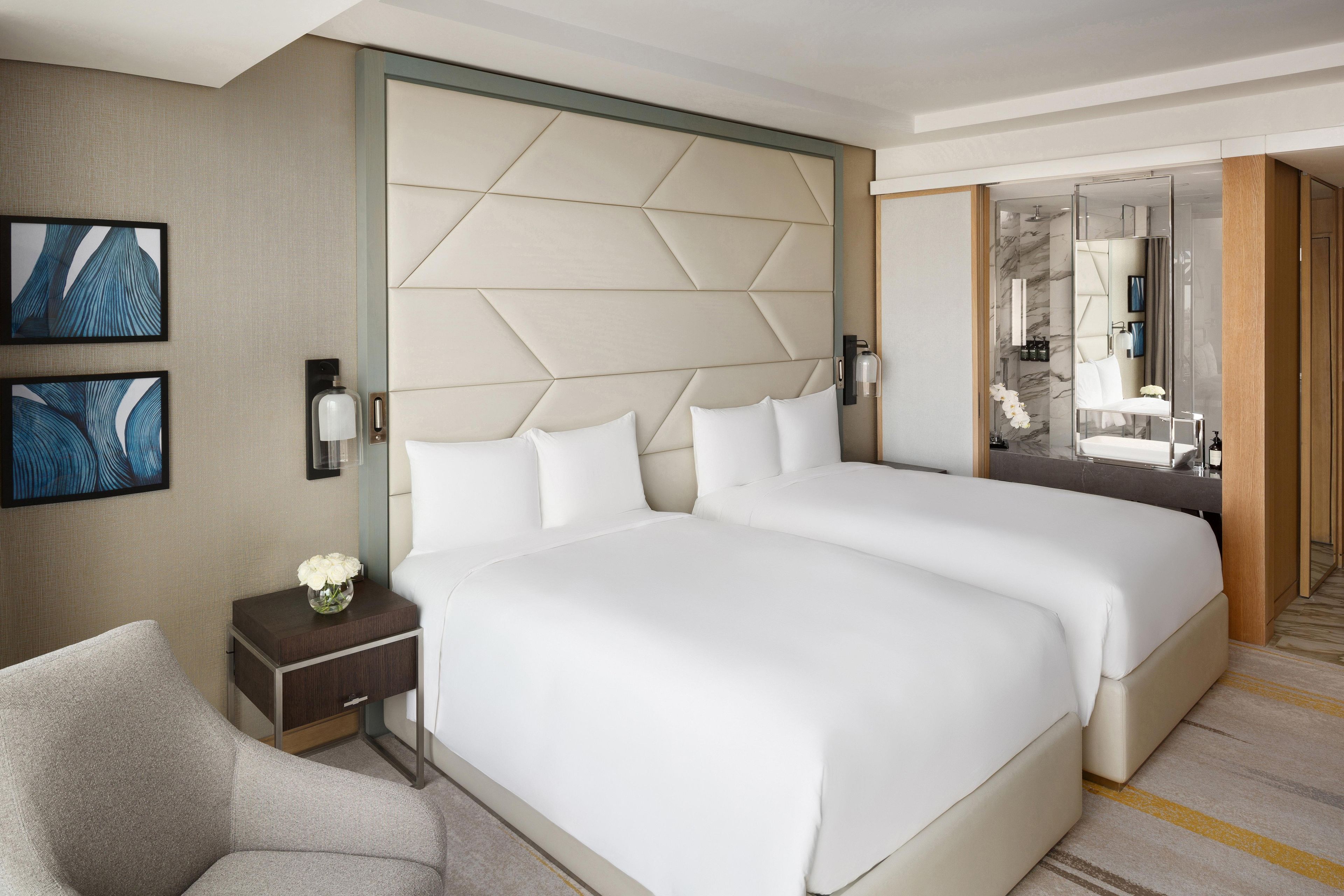 A hotel room with two beds, a nightstand, a chair, a wall-mounted lamp, and a mirror.