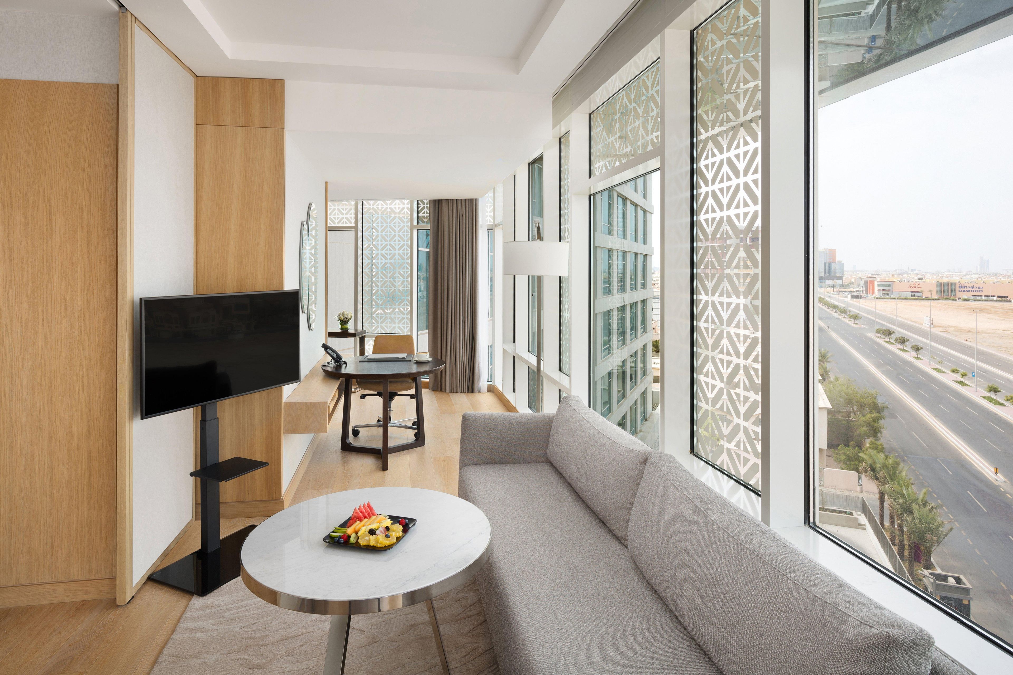A modern hotel room with a large window, a flat-screen TV, a sofa, a small table, and a dining area.
