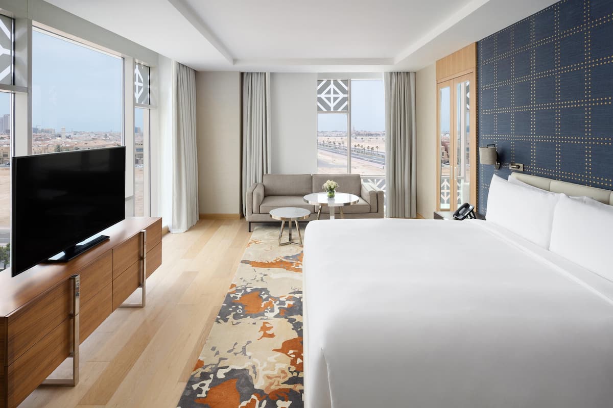 Ein Hotelzimmer mit einem großen Bett, einem Fernseher auf einer Holzkonsole, einer Couch, einem kleinen runden Tisch und großen Fenstern mit Blick auf die Stadt.