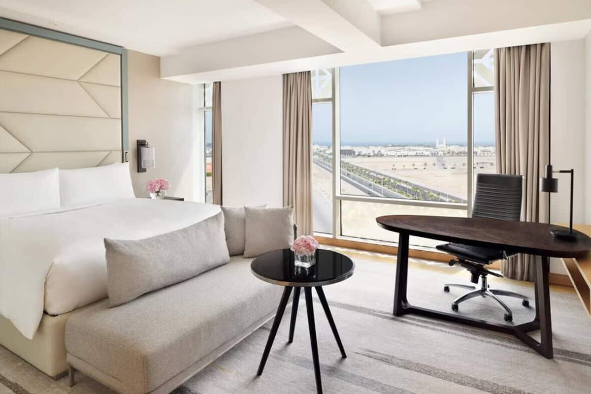 Ein Hotelzimmer mit Bett, Sofa, Schreibtisch, Stuhl und großen Fenstern mit Blick auf die Stadt.