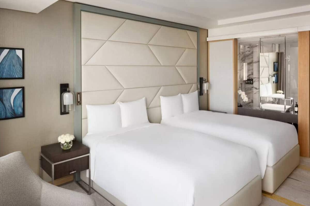 Ein Hotelzimmer mit zwei Betten, einem Nachttisch, einem Sessel und einem durch eine Glastrennwand abgetrennten Badezimmer.