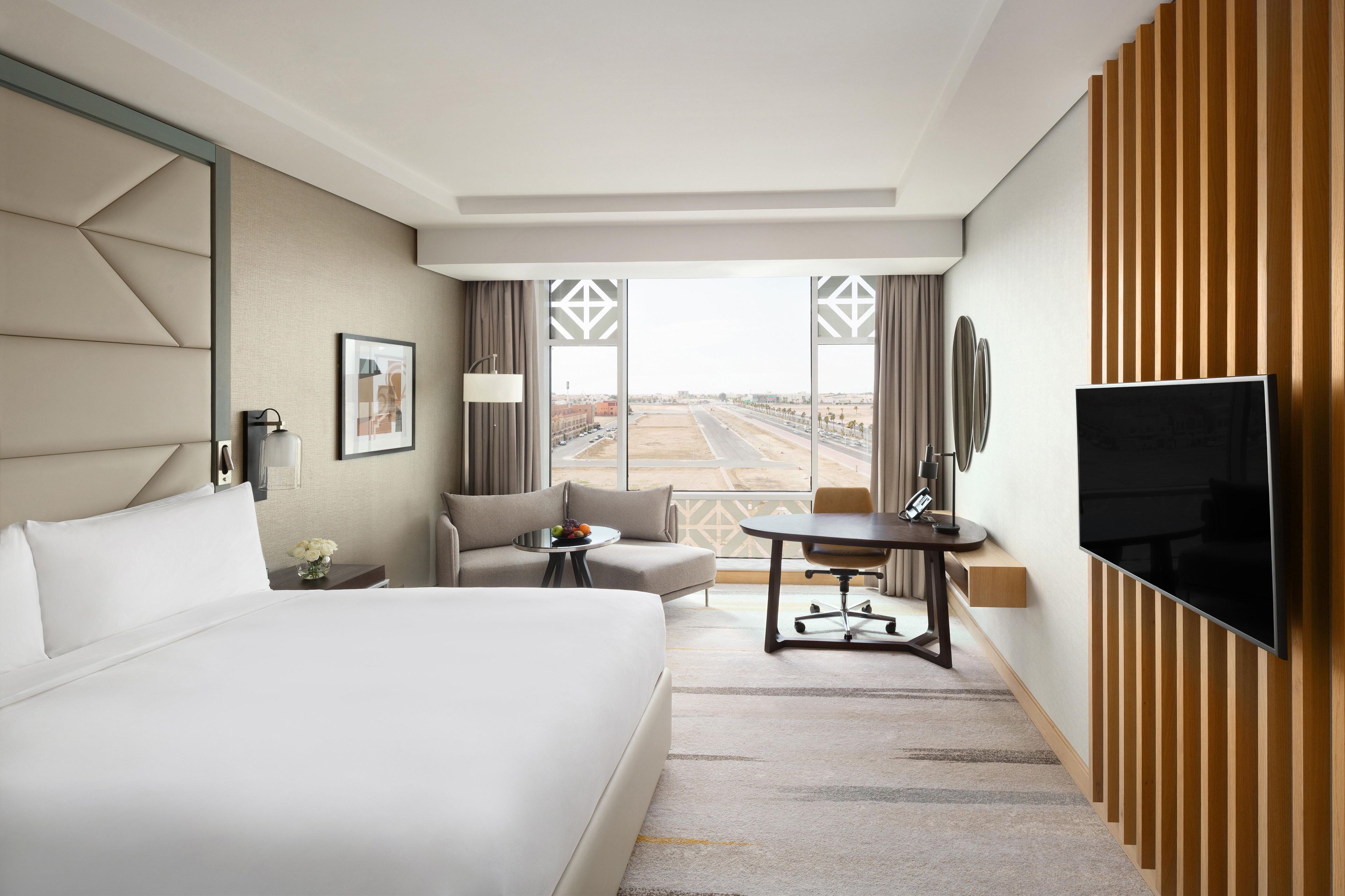 Habitación de hotel con una cama grande, un escritorio, una silla, un televisor y vistas al aeropuerto.