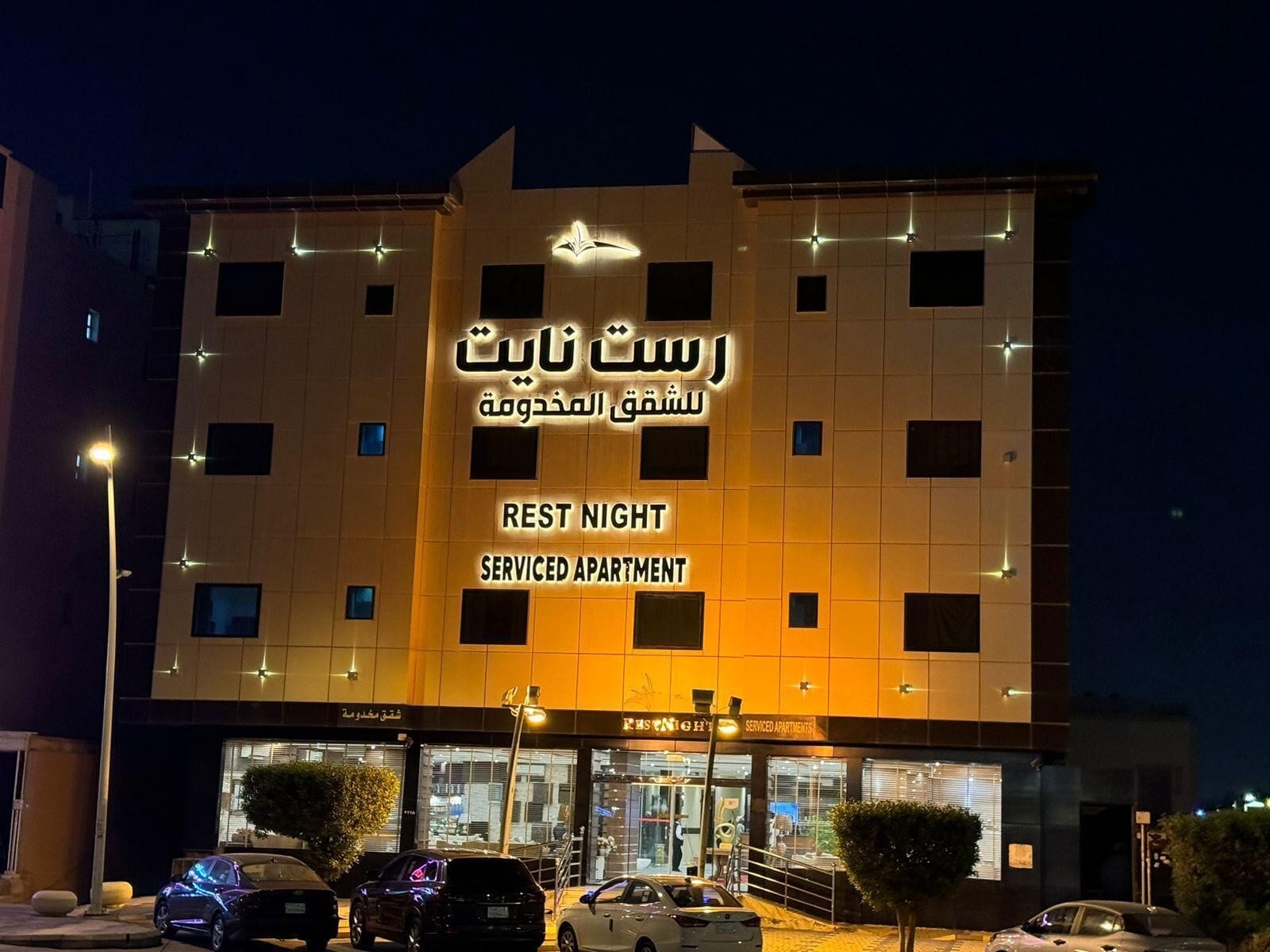 Un edificio con letreros iluminados que indican 'Rest Night Serviced Apartments' y varios coches estacionados en la parte delantera.