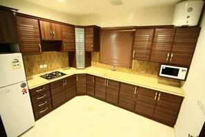 A compact kitchen with dark wooden cabinets, a white refrigerator, a gas stove, and a microwave. - Rest Night Serviced Apartments - AL Nafal (Riyadh)