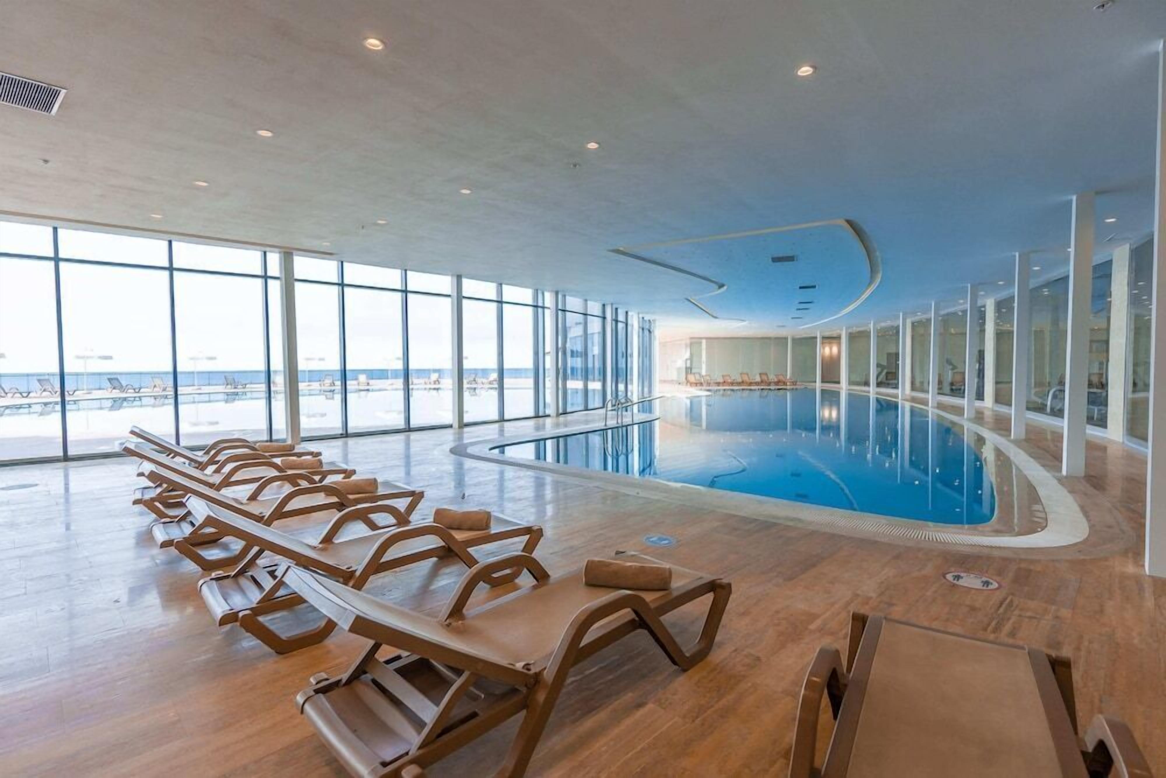 Une piscine intérieure dotée de chaises longues et de grandes fenêtres offrant une vue sur la mer.