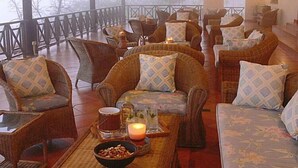 A covered outdoor seating area with wicker furniture and cushions, a table with a candle and snacks, and a view of trees through the railing. - Pakamisa Private Game Reserve (Pongola)