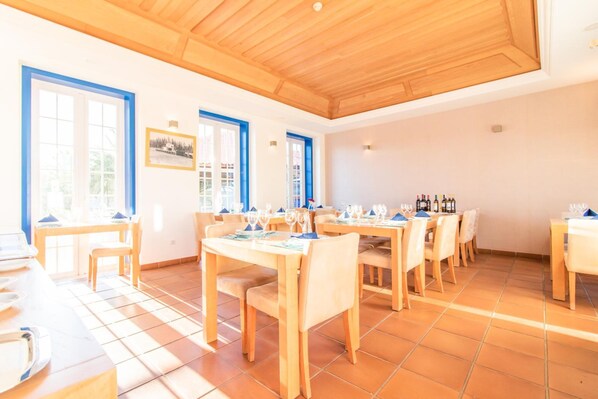 Un comedor con varias mesas y sillas, techo de madera y ventanas grandes con marcos azules.