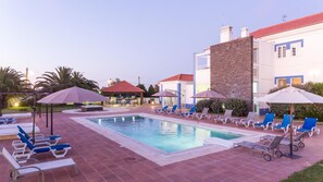 Uma área de piscina com espreguiçadeiras e guarda-sóis, um pequeno bar e um edifício com telhado vermelho.