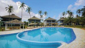 Una piscina con lettini e ombrelloni, circondata da palme e edifici di un hotel.