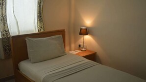 Un letto rifatto con cuscino bianco e testiera in legno, un comodino con una lampada e una finestra con tende a motivi.