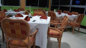 A dining area with round tables covered with white tablecloths, wooden chairs with cushioned seats, and red napkins.