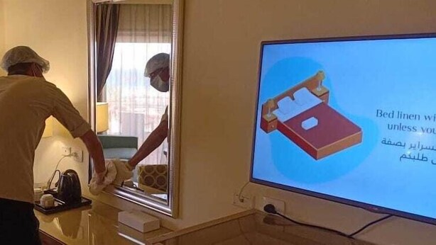 Une personne ajuste un lit dans une chambre d’hôtel, tandis qu’un grand écran affiche des informations sur le linge de lit.