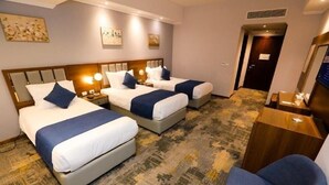 A hotel room with three beds, each with a headboard, bedside tables, and lamps. There is a wooden side table and a chair in the room. - Guard Hotel (Cairo)