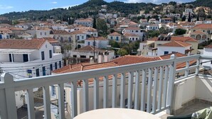A balcony with a round table and chairs, overlooking a hillside town with terracotta-roofed buildings. - Faros (Spetses)
