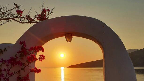 Un chemin en arc de cercle mène à un point de vue pittoresque sur le coucher de soleil sur l’eau, le tout encadré par une structure blanche et des plantes en fleurs.