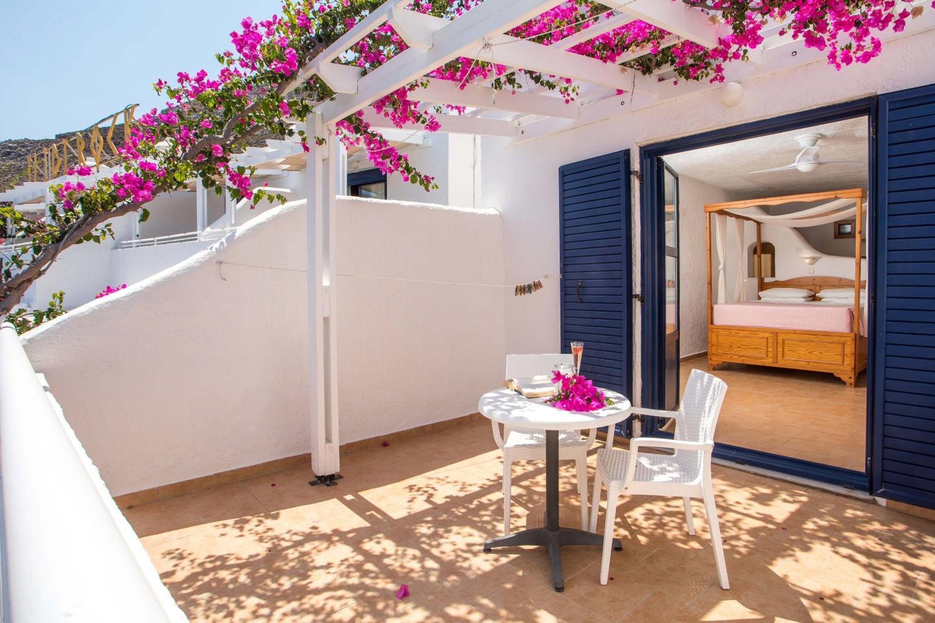 Een balkon met een witte tafel en stoelen, een bed met blauwe luiken, en roze bougainvillea bloemen.
