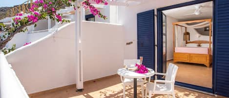 Un balcón con una mesa y sillas blancas, una cama con contraventanas azules y flores de buganvilla rosadas.