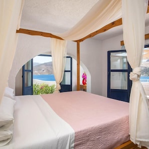 A bedroom with a bed, curtains, and a view of the sea. - Ilidi Rock Apartments (Tilos Island)