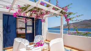 Un balcon agrémenté de meubles en osier blanc et de fleurs roses, offrant une vue sur une mer bleue et un paysage montagneux.