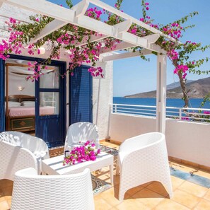 A balcony with white wicker furniture, a bed with a blue door, and pink bougainvillea flowers. - Ilidi Rock Apartments (Tilos Island)