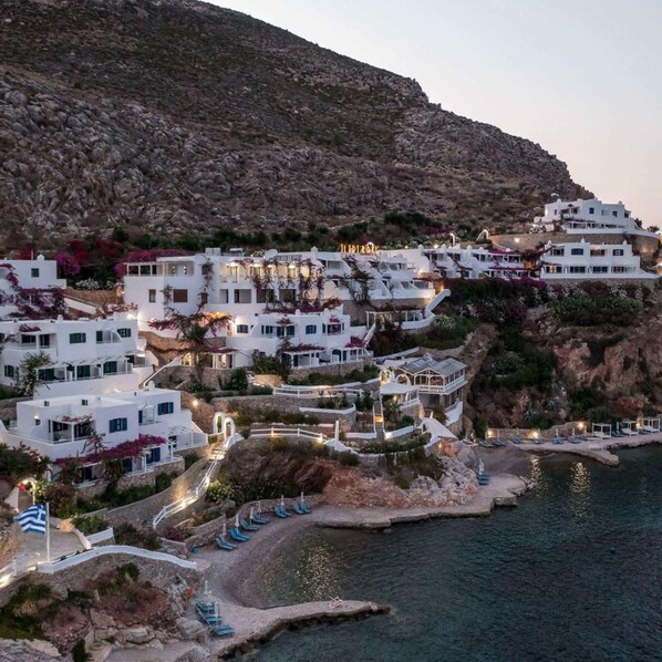 A coastal village with white buildings, a rocky hillside, and a beach with lounge chairs. - Ilidi Rock Apartments (Tilos Island)