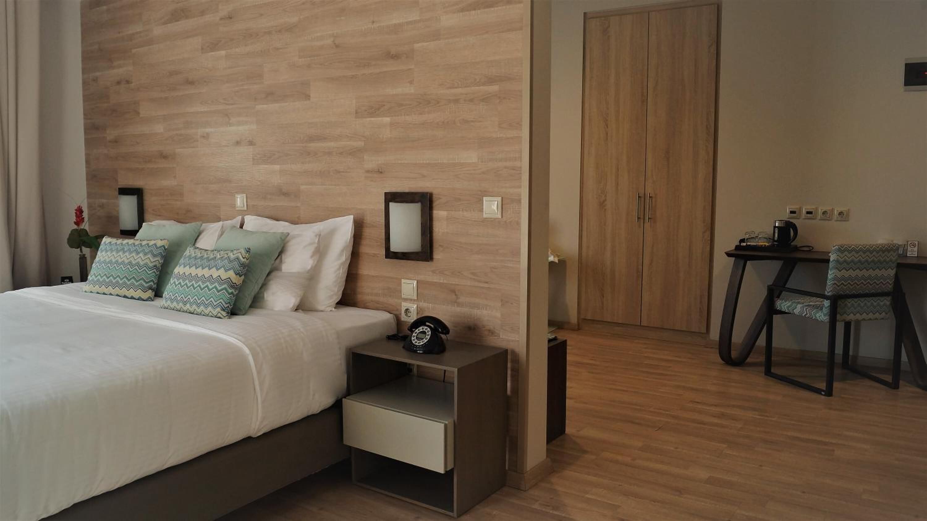 Une chambre d’hôtel avec une tête de lit en bois, un lit avec du linge de maison blanc, une table de chevet avec un téléphone et un bureau avec une chaise.