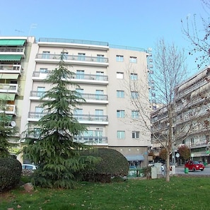 A multi-story building with balconies, a green tree, and a hedge in the foreground. - Park (Larissa)