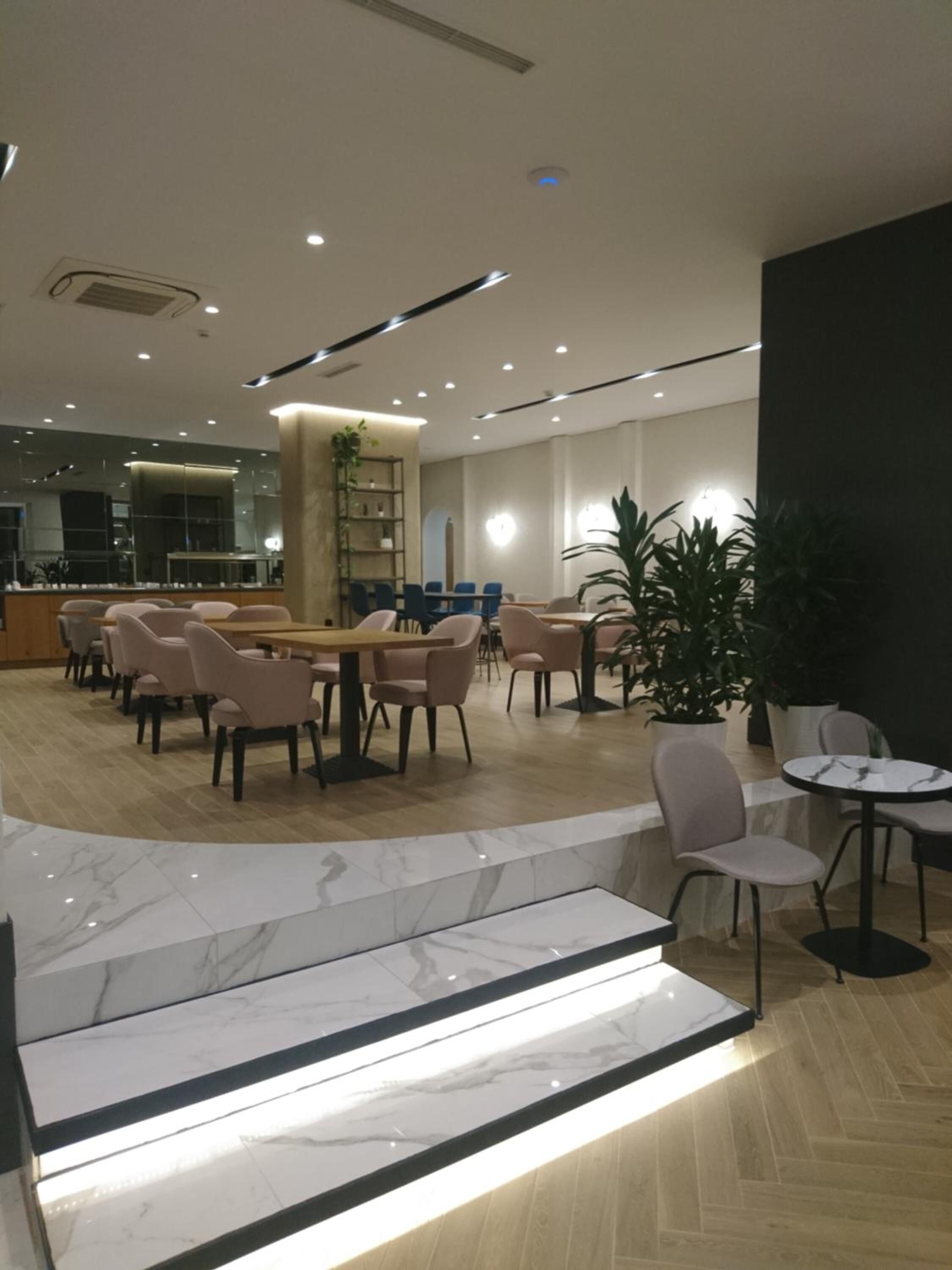 A modern hotel lobby with marble flooring, a central seating area with tables and chairs, and a large potted plant.