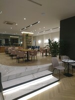 Un moderno vestíbulo de hotel con pisos de mármol, un área de estar central con mesas y sillas, y una planta grande en maceta.