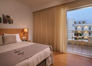 Habitació d'hotel amb llit, una taula de nit, una làmpada, una finestra amb cortines i un balcó amb taula i cadires.