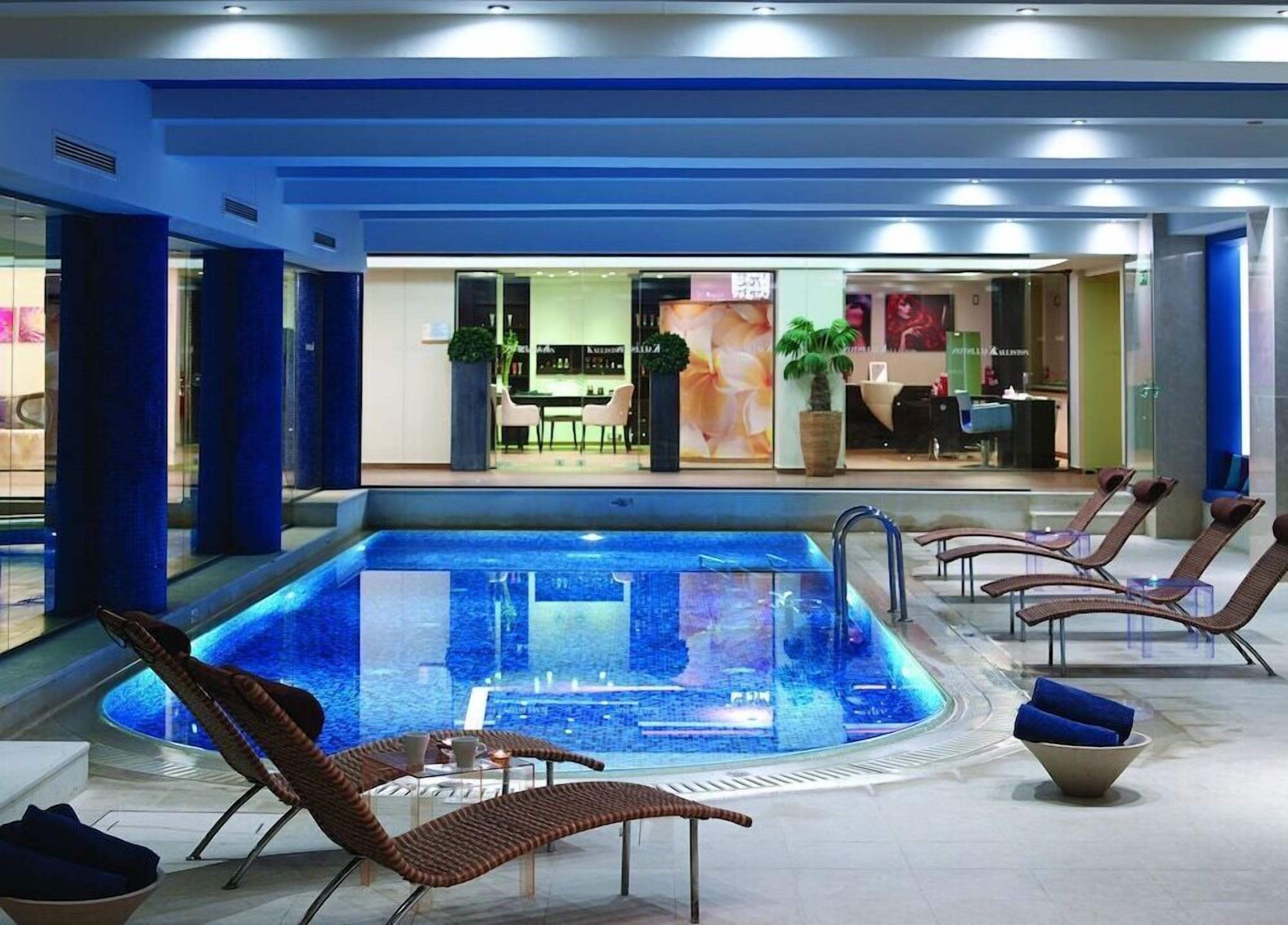 A modern indoor pool area with lounge chairs and a restaurant in the background.